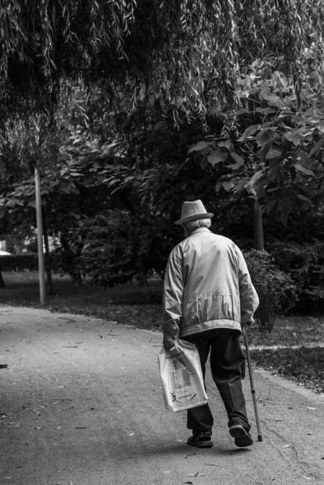 grayscale back view photo of elderly man with cane walking on dirt road